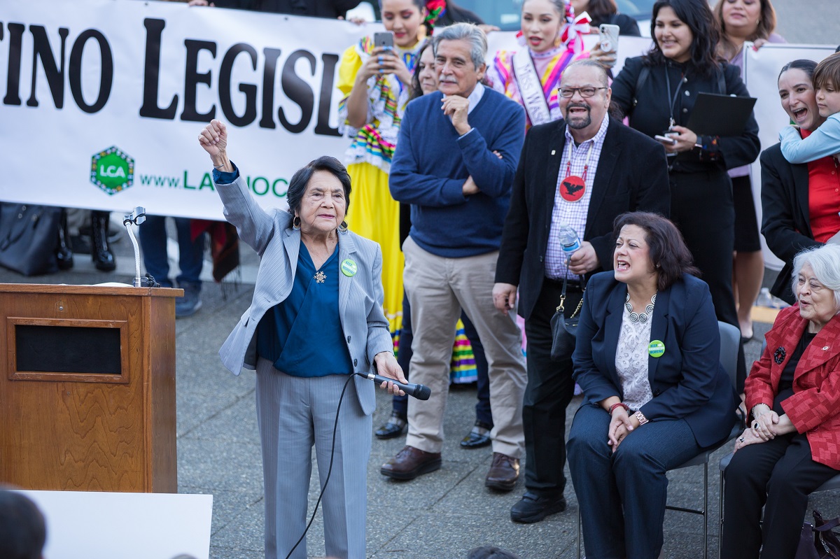 Dolores Huerta stands to the right of a podium, microphone held at her left side, and right fist raised in the air. Next to and behind her colorfully dressed people are gathered and appear to be shouting in support. Part of a Latino Legislative Day sign is visible in the background.