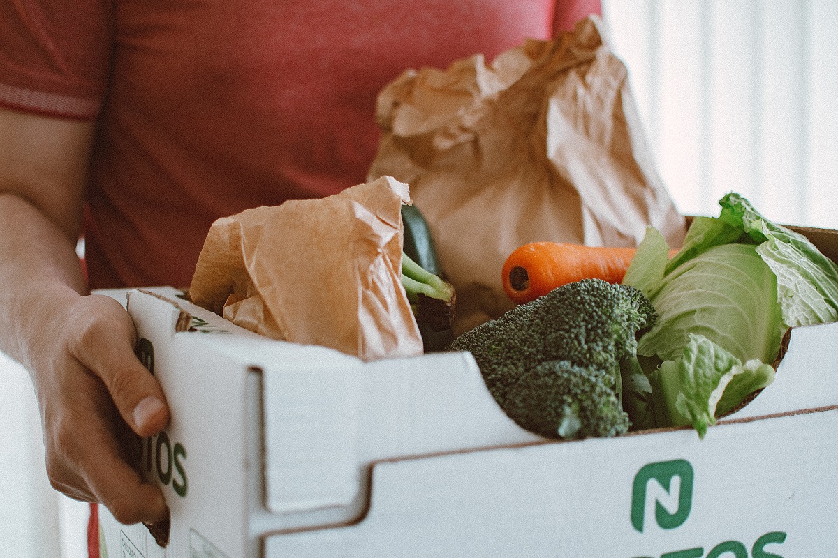 Person holding box full of food. Broccoli, carrots, and lettuce and two brown paper bags are visible in the box.