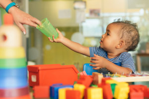 Small child surrounded by colorful toys holds out a transparent green cup being grasped by an adult hand. 