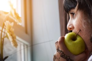 Child looking out a window while biting into a green apple. 