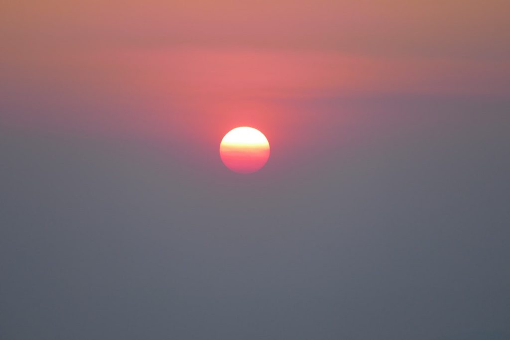 Photo: top third shows red sky, bottom two thirds shows gray smoke. The sun is a small orange and red circle in the middle, partially covered by the smoke.