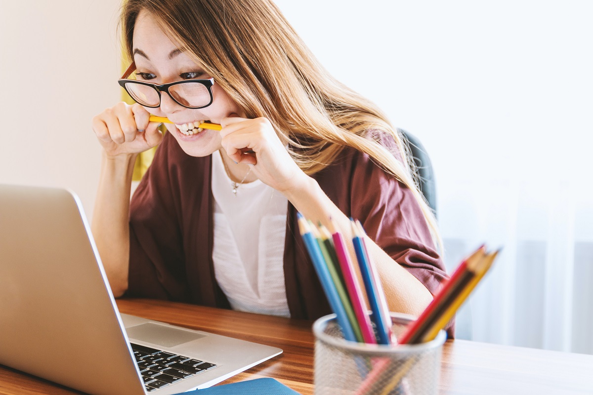 A young woman sits in front of a laptop computer holding a yellow pencil by both ends with her hands, biting down on the pencil in apparent frustration.