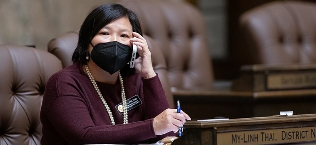 Rep. My-Linh Thai speaks on the phone while wearing a mask at her desk in the House of Representatives chambers in Olympia